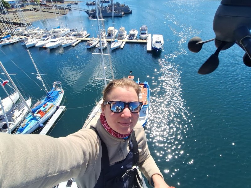 Person on a boat mast taking a selfie, overlooking a marina with several yachts.