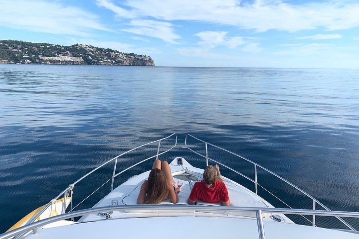 Two people relax on a yacht's deck overlooking calm sea and distant coastline.