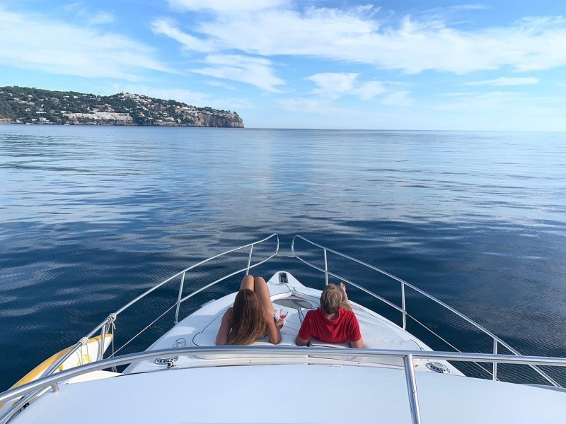 Two people relax on a yacht's deck overlooking calm sea and distant coastline.