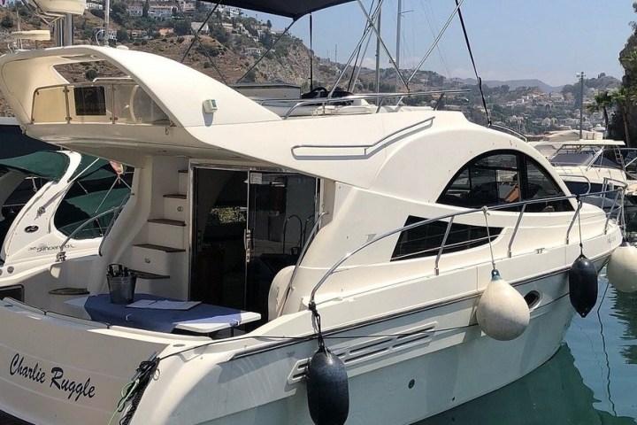 White motor yacht docked in a marina with hills in the background.