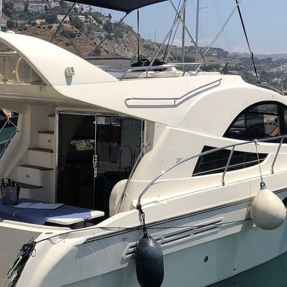 White motor yacht docked in a marina with hills in the background.
