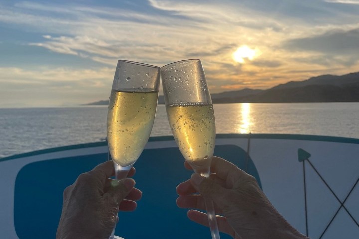 Two hands clinking champagne glasses on a boat at sunset.