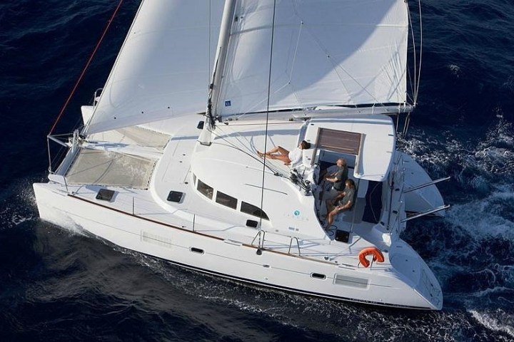 Aerial view of a white catamaran sailing with two people on deck in deep blue water.