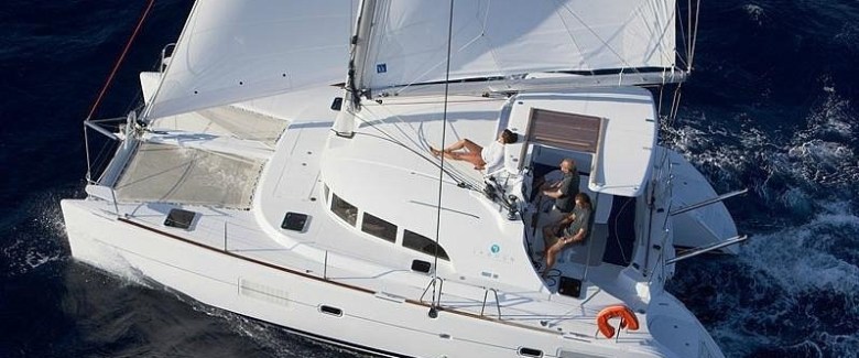 Aerial view of a white catamaran sailing with two people on deck in deep blue water.