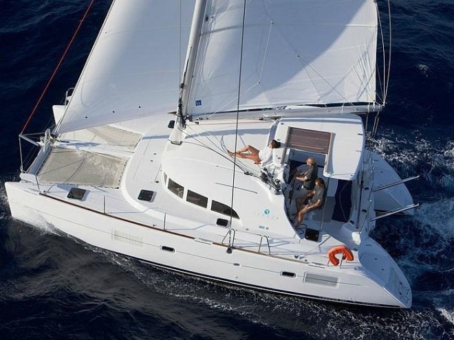 Aerial view of a white catamaran sailing with two people on deck in deep blue water.