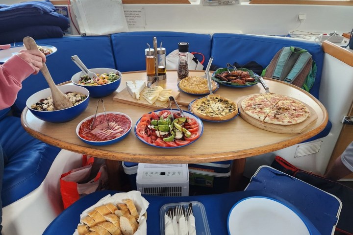 Table with assorted dishes including salads, pizza, cheese, meats, and a loaf of bread on a yacht.