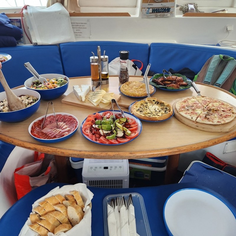 Table with assorted dishes including salads, pizza, cheese, meats, and a loaf of bread on a yacht.