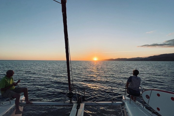Two people on a catamaran watching a sunset over the ocean with mountains in the distance.