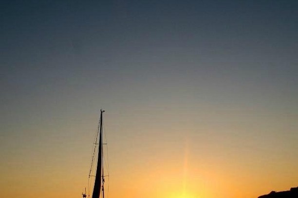 Sailboat on calm water at sunrise with an orange sky and distant cliffs.