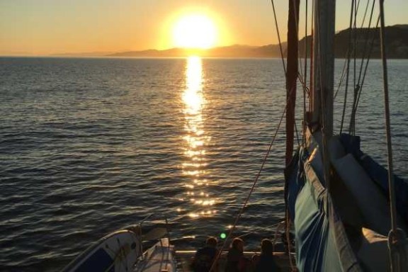 Sunset over ocean with sailboat in foreground, silhouetted figures on deck.