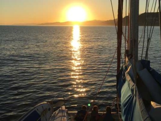 Sunset over ocean with sailboat in foreground, silhouetted figures on deck.