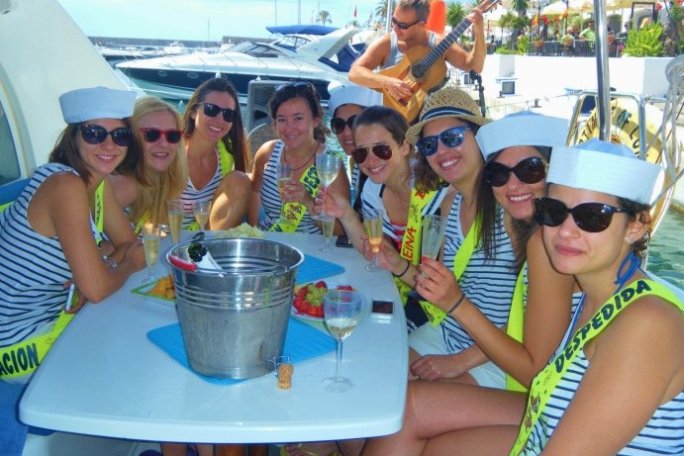 Group of women in sailor hats on a boat, holding drinks, with fruit and a musician in the background.