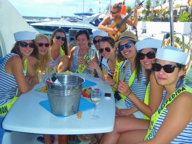 Group of women in sailor hats on a boat, holding drinks, with fruit and a musician in the background.