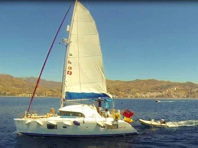 Sailing catamaran with white sails on calm sea, people on deck, mountains and city in background.