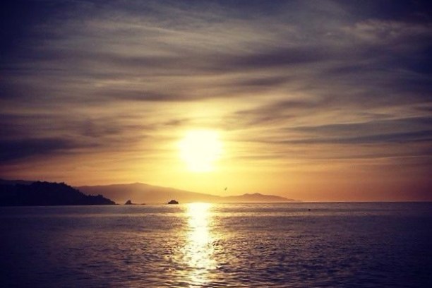 A serene sunset over the ocean with silhouetted hills and a cloudy sky.