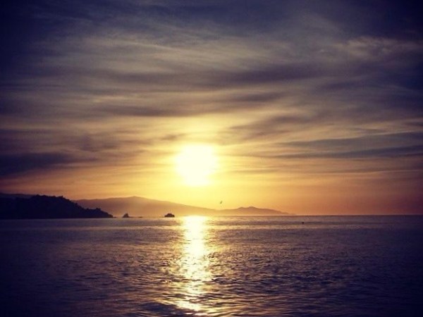A serene sunset over the ocean with silhouetted hills and a cloudy sky.