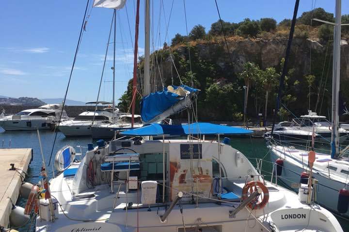 Sailboat named 'Optimist of London VI' docked at a marina with a 'CHARTER' flag and nearby yachts.
