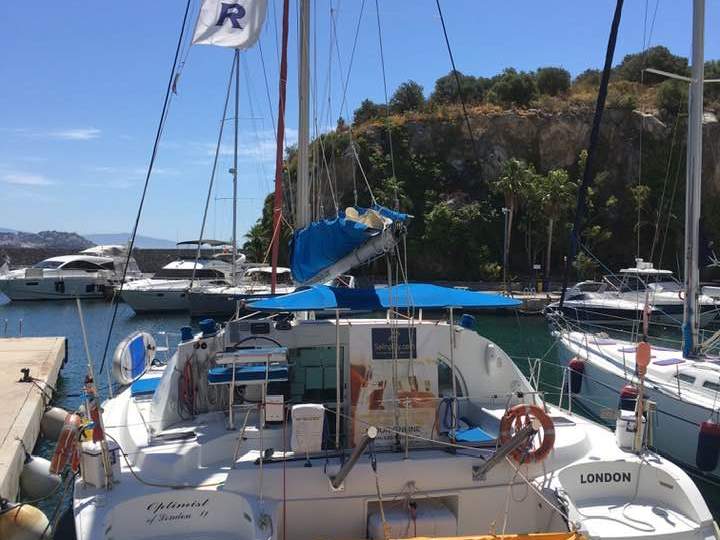 Sailboat named 'Optimist of London VI' docked at a marina with a 'CHARTER' flag and nearby yachts.