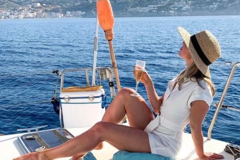Woman relaxing on a yacht, wearing a hat and holding a glass of wine, with mountains in the background.