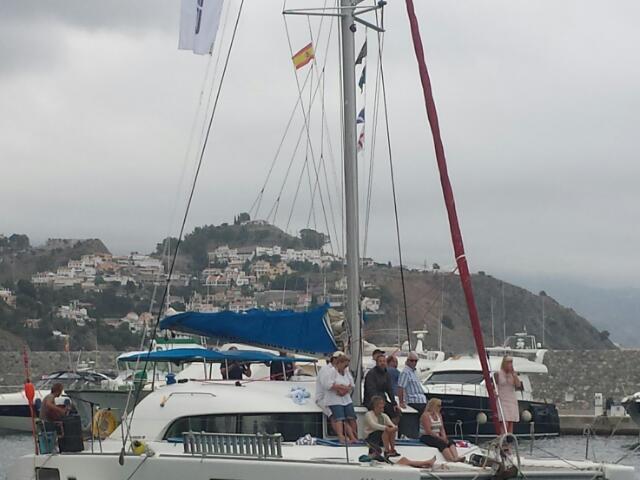 People on a sailboat in a marina with hillside houses in the background.