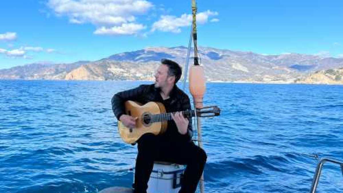Man with guitar sitting on a boat, ocean and mountains in the background.