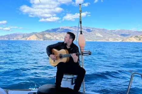 Man with guitar sitting on a boat, ocean and mountains in the background.