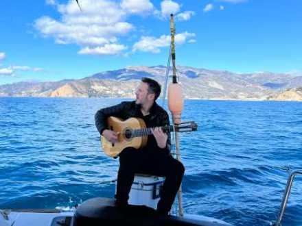 Man with guitar sitting on a boat, ocean and mountains in the background.