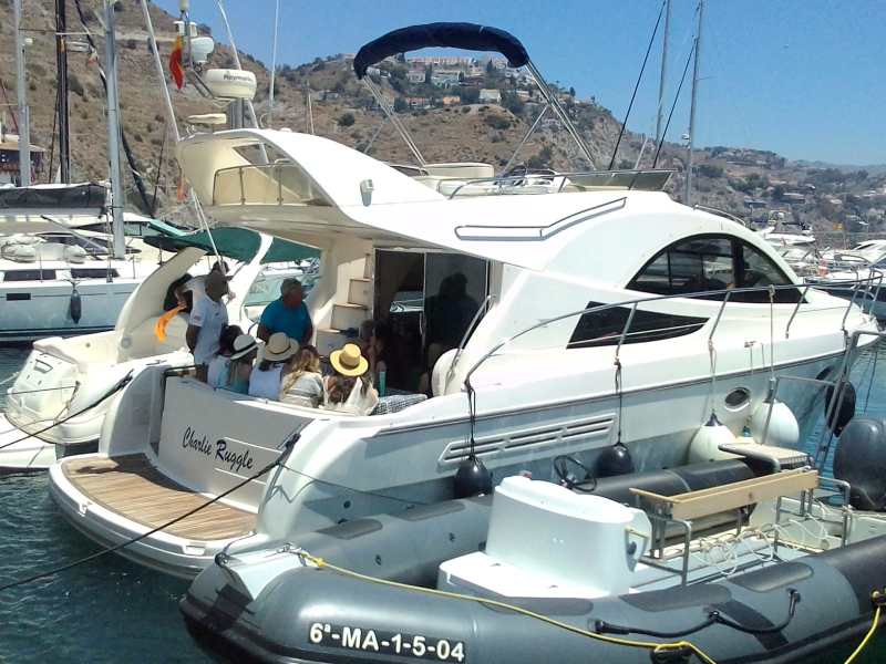 Group of people on a docked white yacht with hills and houses in the background.