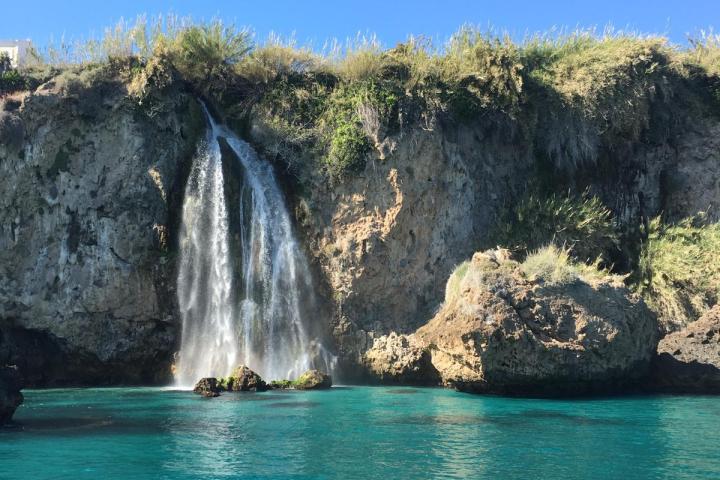 Waterfall cascading over rocky cliff into turquoise sea under clear blue sky.