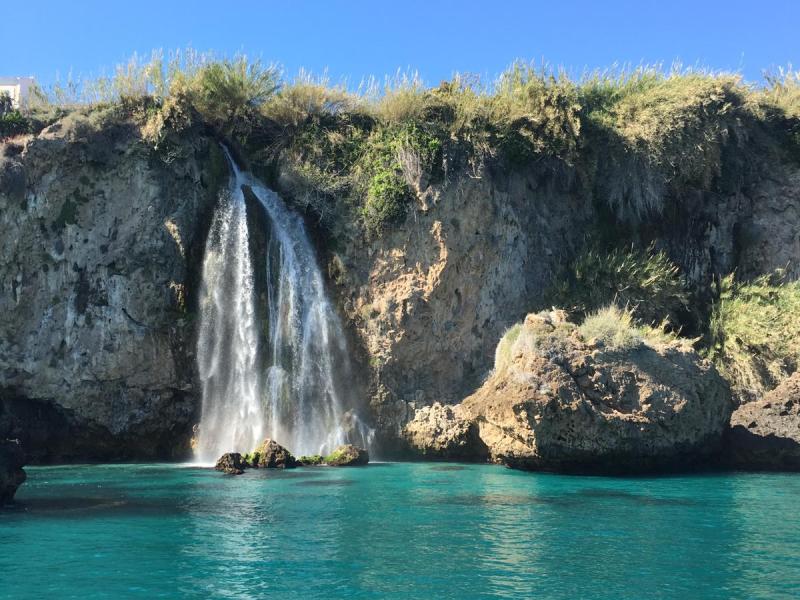 Waterfall cascading over rocky cliff into turquoise sea under clear blue sky.