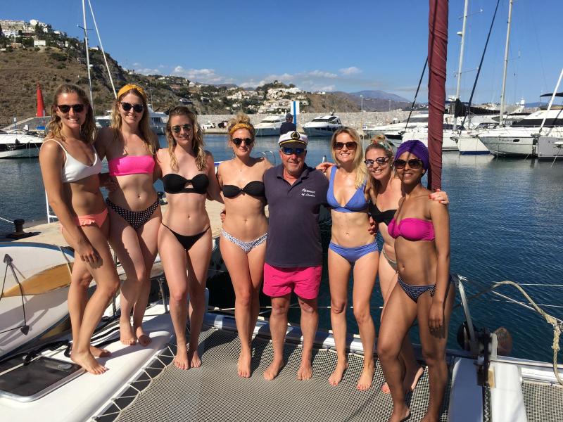 Group of people in swimwear posing on a yacht with a marina and hills in the background.