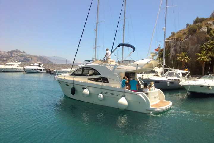 Motor yacht with people aboard in a marina surrounded by other boats and hills.