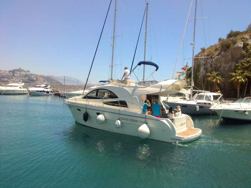 Motor yacht with people aboard in a marina surrounded by other boats and hills.