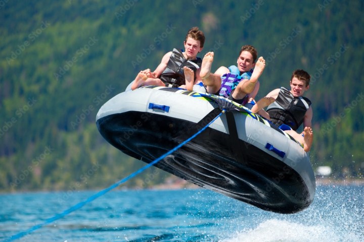 Three people wearing life jackets ride an inflatable tube over water, with mountains in the background.
