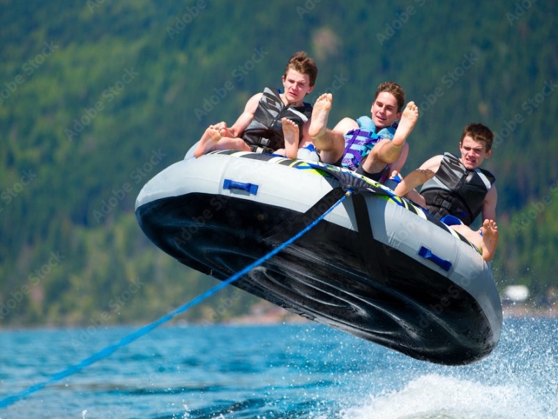 Three people wearing life jackets ride an inflatable tube over water, with mountains in the background.