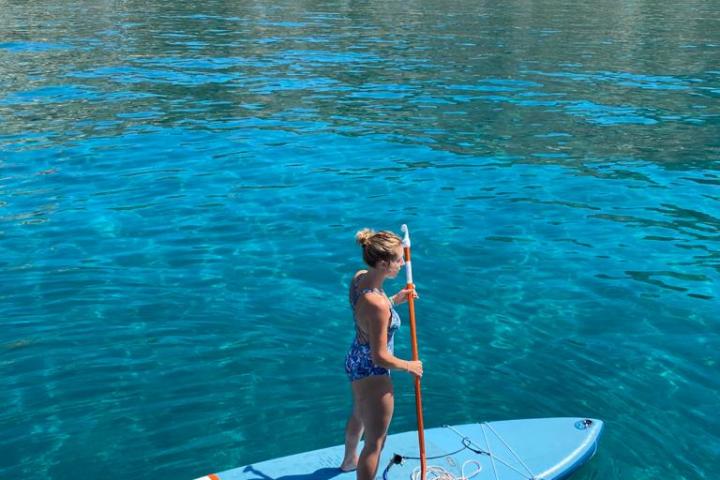 Person paddleboarding on clear blue water near a scenic beach and hills.