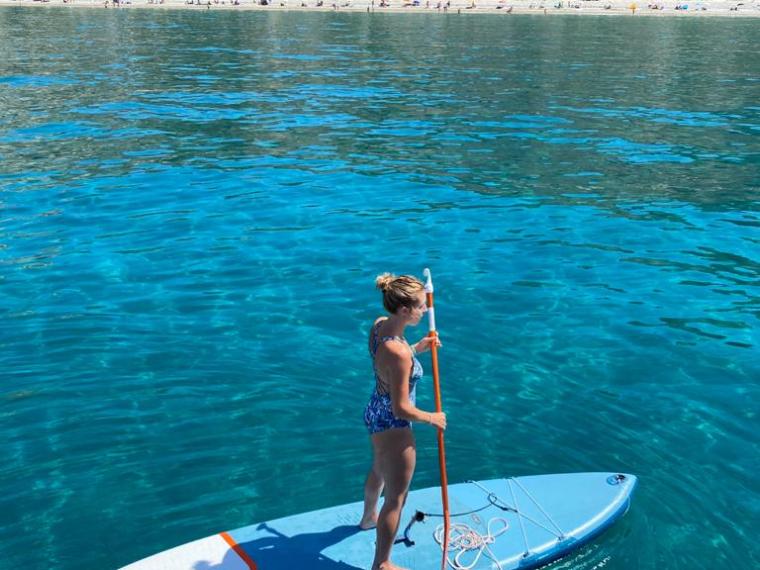 Person paddleboarding on clear blue water near a scenic beach and hills.