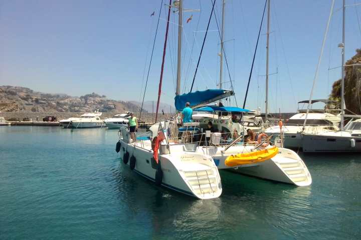 People on a sailboat docked in a marina with other boats under a clear sky.
