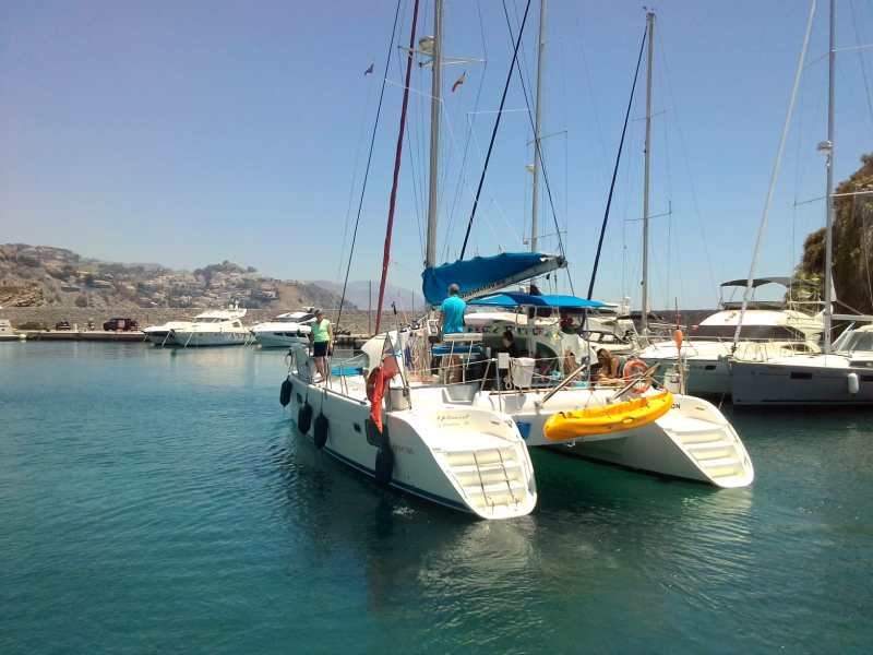 People on a sailboat docked in a marina with other boats under a clear sky.
