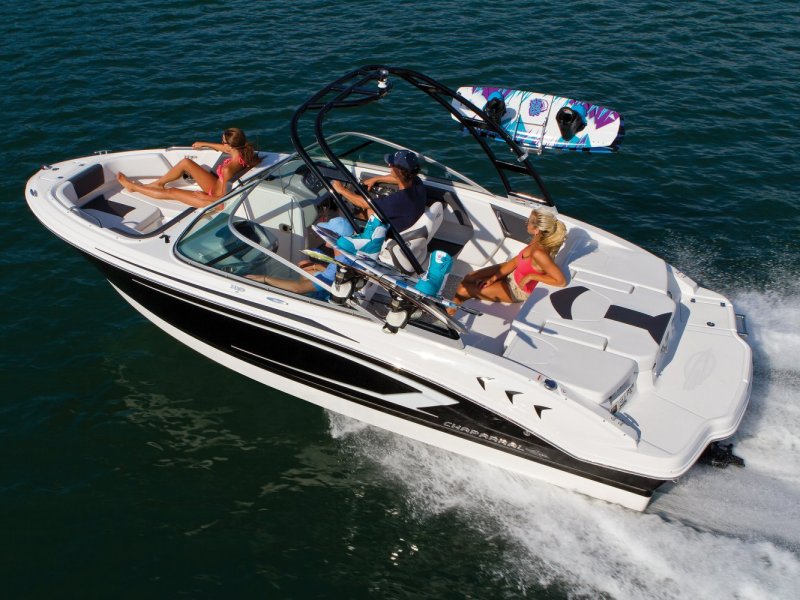Speedboat with four people on a sunny day, one wakeboard on rack, moving through water.