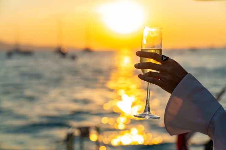 Hand holding a champagne flute against a sunset over a calm sea.
