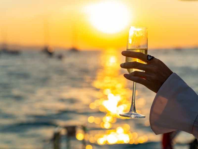 Hand holding a champagne flute against a sunset over a calm sea.