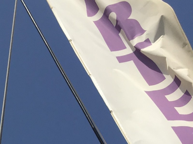 White flag with purple letters waving on a pole against a clear blue sky.