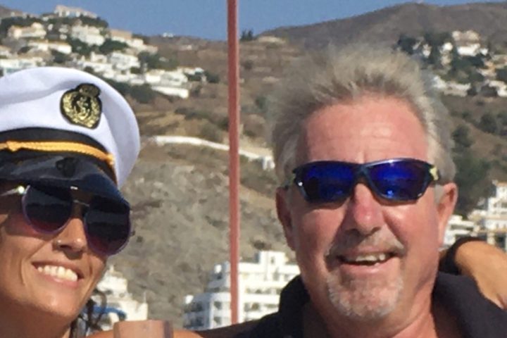 Two people on a boat, one in a captain's hat holding a glass, both wearing sunglasses, with hills in the background.