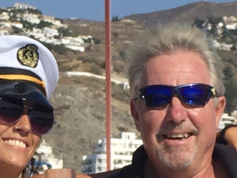 Two people on a boat, one in a captain's hat holding a glass, both wearing sunglasses, with hills in the background.