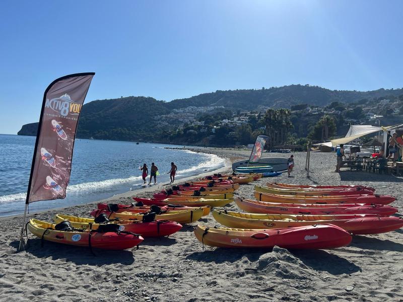 Colorful kayaks lined up on a beach with a nearby flag and people walking by the sea.