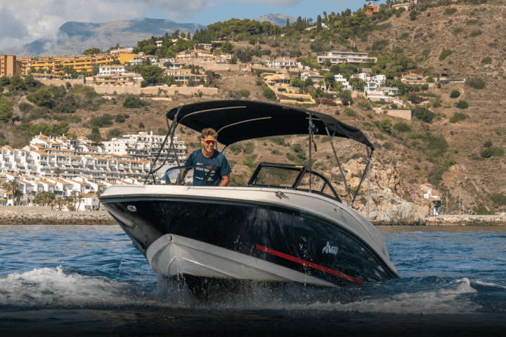 Man driving a motorboat on water with hillside houses in the background.