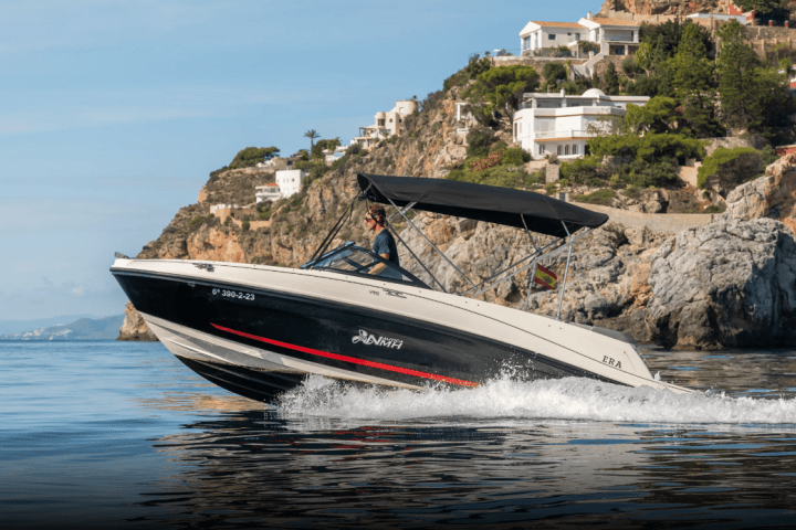 Motorboat with canopy cruising near rocky coastline with houses above.