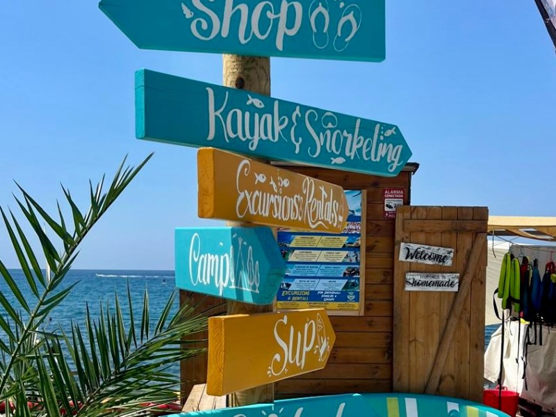 Colorful wooden signpost with ocean activities near a beach hut.