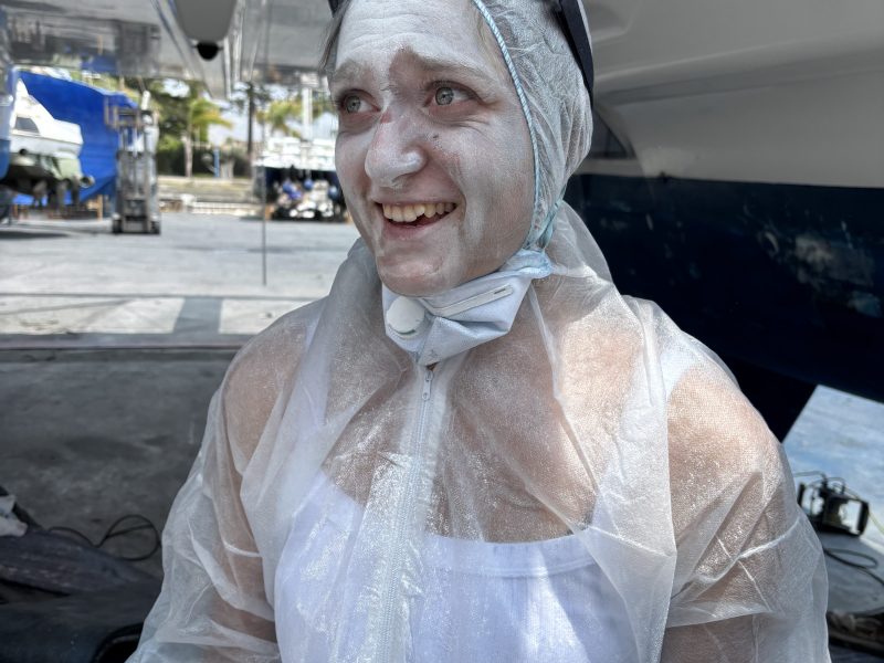 Person in white protective suit with goggles, face covered in white powder, smiling in boat workshop.